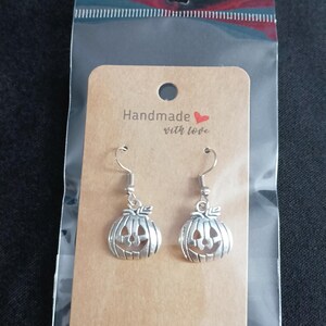 May include: Silver pumpkin earrings with a carved jack-o'-lantern face, presented on a brown card with the text "Handmade with love." The earrings are packaged in a clear plastic bag, ideal for Halloween.