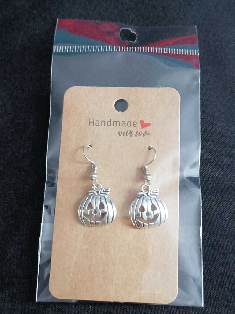 May include: Silver pumpkin earrings with a carved jack-o'-lantern face, presented on a brown card with the text "Handmade with love." The earrings are packaged in a clear plastic bag, ideal for Halloween.