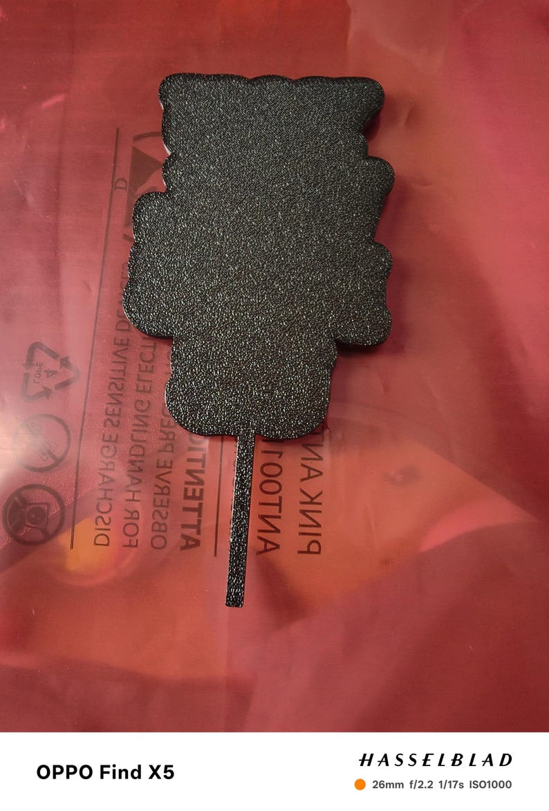 May include: A black, textured, decorative item with a unique shape, possibly a pendant or ornament. The item is set against a red background. The image was taken with an OPPO Find X5 camera.
