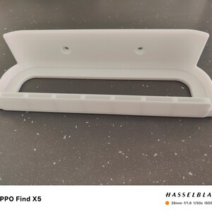 May include: A white, wall-mounted shelf with a curved handle design. The shelf has a rectangular opening and several small rectangular slots along the bottom edge. Two holes are visible near the top. The shelf is made of a solid, opaque material.