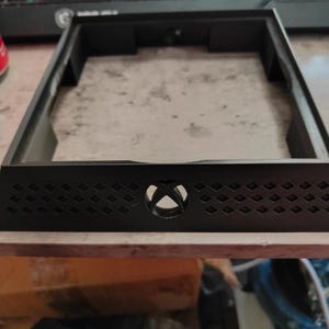 May include: A black, rectangular frame with an Xbox logo in the center. The frame has a diamond-patterned design along the front edge and is likely a component for a gaming console or related device.