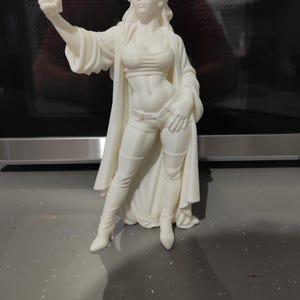 K&ouml;nnte beinhalten: Eine wei&szlig;e Statue der Freiheitsstatue, die ein Selfie mit einem Telefon macht. Die Statue tr&auml;gt ein bauchfreies Oberteil, Shorts und Stiefel. Die Statue hat eine Krone mit Zacken. Die Statue steht auf einer grauen Oberfl&auml;che.