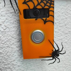 May include: Orange doorbell cover decorated with black spiders and a spiderweb design. The doorbell has a silver button and a camera lens. The cover is mounted on a textured white wall.