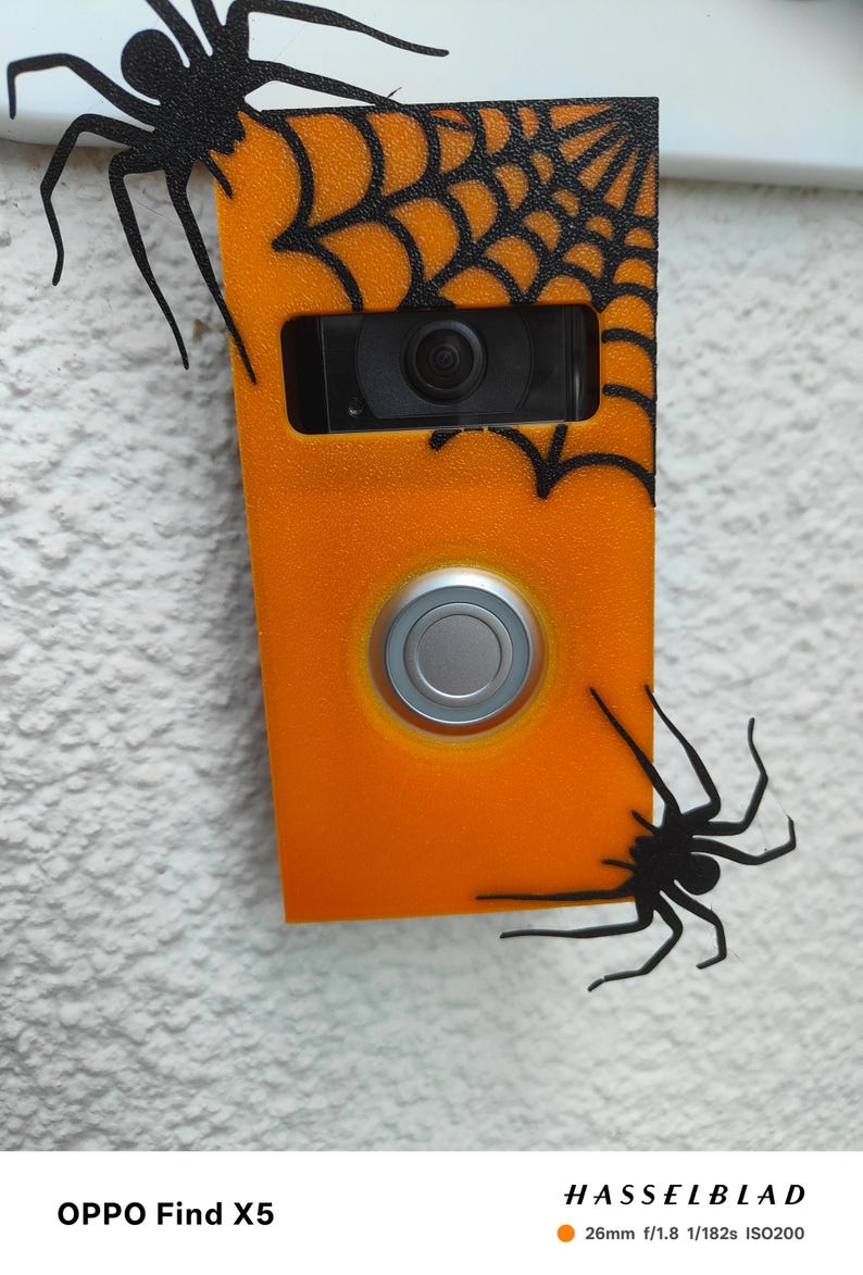 May include: Orange doorbell cover decorated with black spiders and a spiderweb design. The doorbell has a silver button and a camera lens. The cover is mounted on a textured white wall.