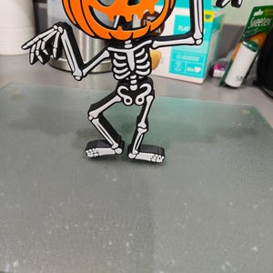 May include: A Halloween-themed decoration featuring a pumpkin head with a skeleton body. The pumpkin is orange with black facial features and a black stem. The skeleton is black with white bone details. The figure is standing and pointing with its arms.