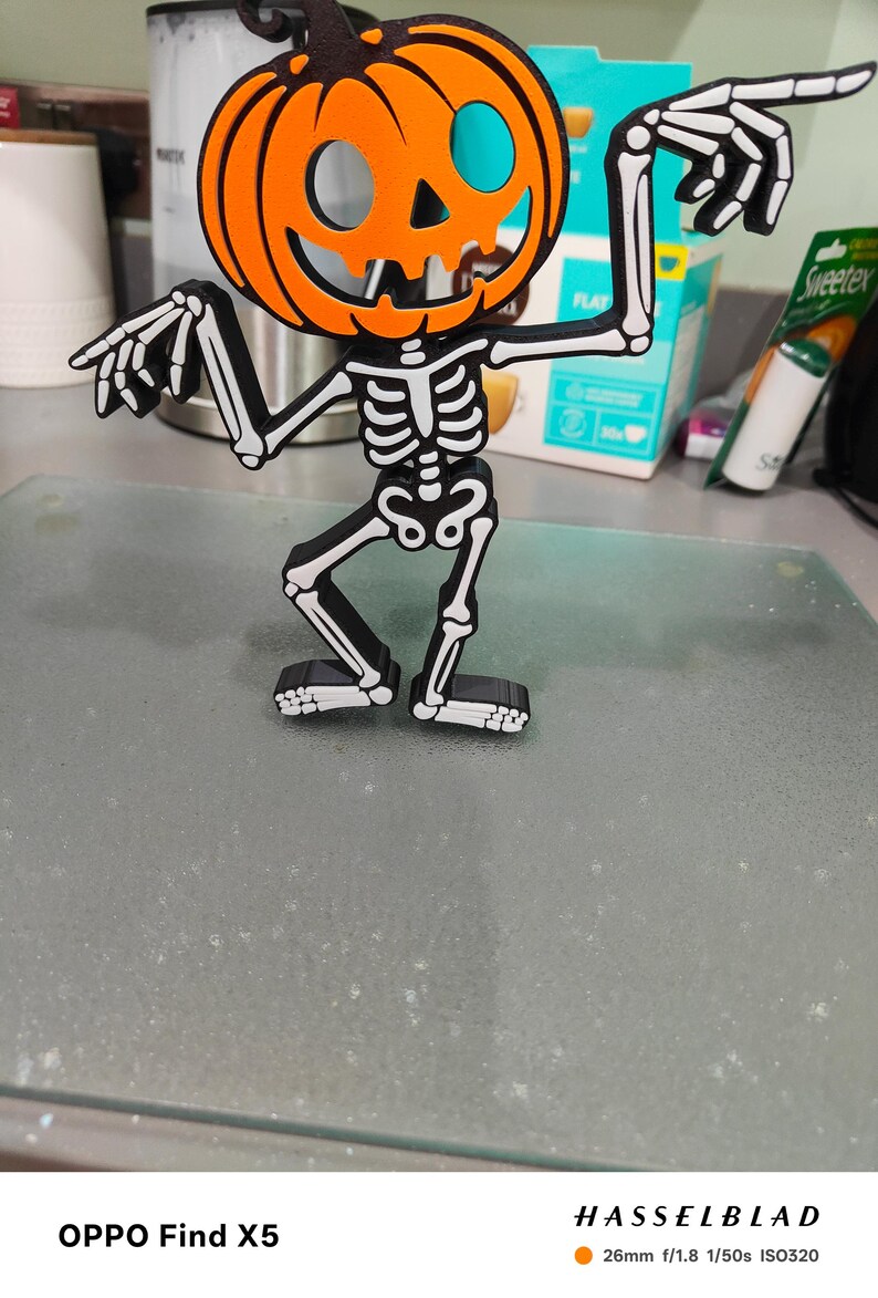 May include: A Halloween-themed decoration featuring a pumpkin head with a skeleton body. The pumpkin is orange with black facial features and a black stem. The skeleton is black with white bone details. The figure is standing and pointing with its arms.