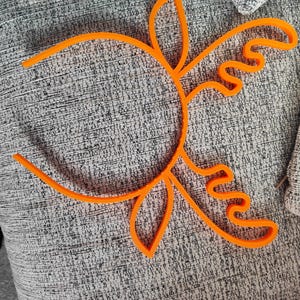 Puede incluir: Dise&ntilde;o floral abstracto de color naranja brillante. Se ve el contorno de una flor o un sol. El objeto est&aacute; hecho de un material s&oacute;lido y se encuentra sobre una superficie gris texturizada. La imagen est&aacute; bien iluminada.