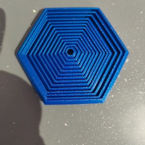 May include: A bright blue, hexagonal coaster with a concentric pattern. The coaster has a series of nested, six-sided shapes, creating a textured surface. The central hole adds a unique detail to this functional item.