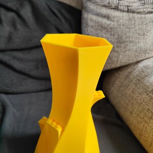 May include: A bright yellow, geometric vase with a unique, twisted design. The vase features small, rectangular blocks on the sides, adding a playful touch. The vase is made of a solid material and has a modern aesthetic.