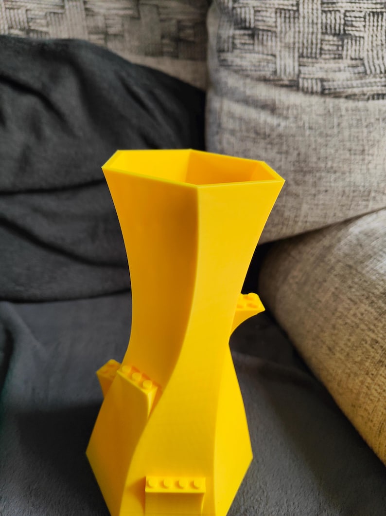 May include: A bright yellow, geometric vase with a unique, twisted design. The vase features small, rectangular blocks on the sides, adding a playful touch. The vase is made of a solid material and has a modern aesthetic.