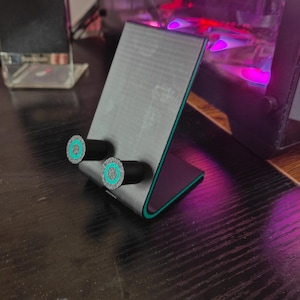 K&ouml;nnte beinhalten: Ein grauer Telefonst&auml;nder mit schwarzem Sockel und zwei schwarzen zylindrischen Halterungen mit t&uuml;rkisfarbenen und grauen kreisf&ouml;rmigen Designs. Der St&auml;nder hat eine lila und gr&uuml;ne Umrandung. Der Text "OPPO Find X5" steht unten.