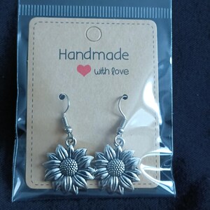 May include: A pair of silver-colored sunflower earrings with a detailed design, displayed on a brown card that reads "Handmade with love." The earrings are packaged in a clear plastic bag.