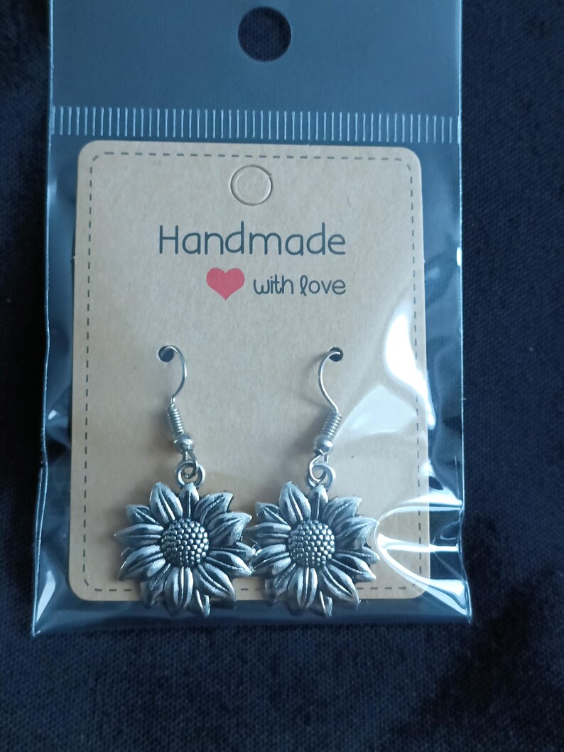 May include: A pair of silver-colored sunflower earrings with a detailed design, displayed on a brown card that reads "Handmade with love." The earrings are packaged in a clear plastic bag.