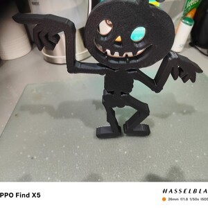 May include: A black Halloween decoration in the shape of a pumpkin-headed skeleton. The figure has outstretched arms with pointing fingers and a smiling jack-o'-lantern face. The decoration is made of a matte black material and stands on a flat base.