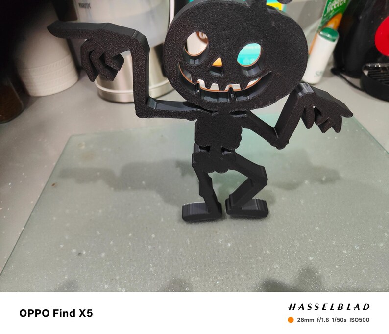 May include: A black Halloween decoration in the shape of a pumpkin-headed skeleton. The figure has outstretched arms with pointing fingers and a smiling jack-o'-lantern face. The decoration is made of a matte black material and stands on a flat base.