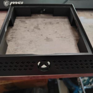 May include: A black rectangular console stand with an Xbox logo and diamond-shaped cutouts. The stand has a raised frame and is designed to hold a gaming console. The stand is made of plastic.