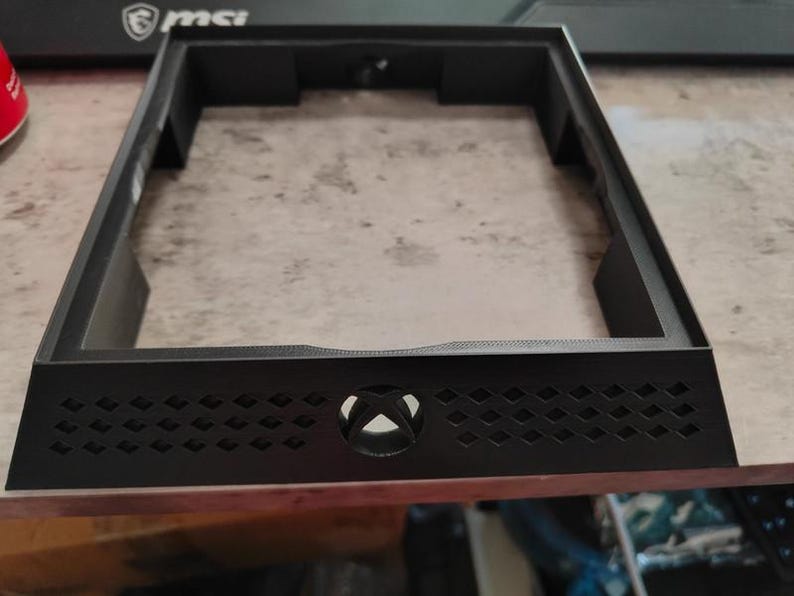 May include: A black rectangular console stand with an Xbox logo and diamond-shaped cutouts. The stand has a raised frame and is designed to hold a gaming console. The stand is made of plastic.