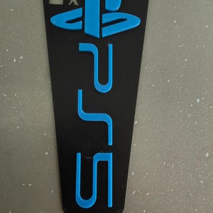 May include: Black rectangular phone case with a blue PlayStation 5 logo and symbols. The case features the PlayStation logo and the letters "PS5" in a vibrant blue color, set against a black background. The case also includes the square, triangle, X, and circle symbols.