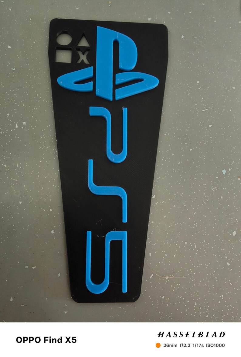 May include: Black rectangular phone case with a blue PlayStation 5 logo and symbols. The case features the PlayStation logo and the letters "PS5" in a vibrant blue color, set against a black background. The case also includes the square, triangle, X, and circle symbols.