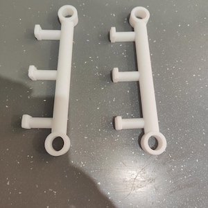 May include: Two white plastic components with a series of perpendicular pegs and a circular opening at each end. The components are identical and appear to be 3D-printed. The image is taken from above.