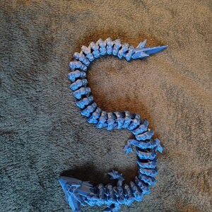 May include: A blue articulated dragon toy, with a segmented body and pointed features. The dragon is made of plastic and is posed on a textured surface. The dragon's body is curved, and it has a tail and head.