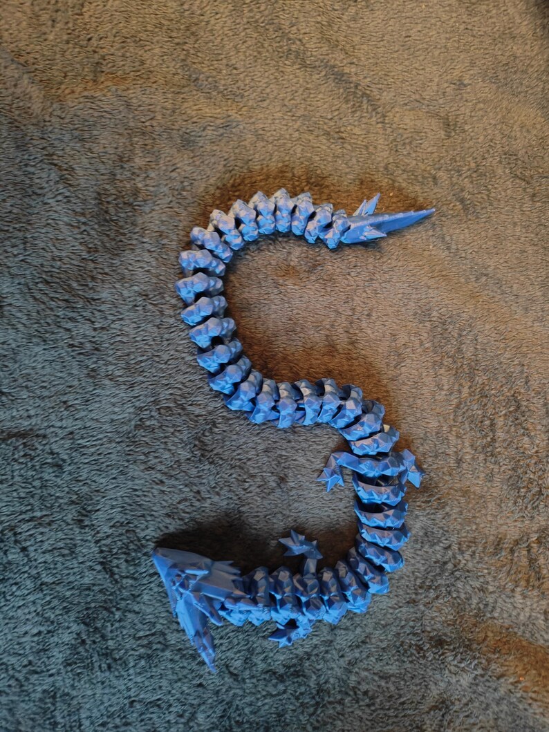 May include: A blue articulated dragon toy, with a segmented body and pointed features. The dragon is made of plastic and is posed on a textured surface. The dragon's body is curved, and it has a tail and head.