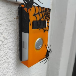 May include: A Halloween-themed doorbell camera with an orange faceplate, black accents, and spider decorations. The doorbell has a silver button and a camera lens. A spider web design is visible on the orange faceplate.