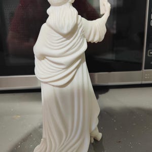 K&ouml;nnte beinhalten: Eine wei&szlig;e Statue der Freiheitsstatue, die ein Selfie mit einem Smartphone macht. Die Statue tr&auml;gt eine stachelige Krone und drapierte Gew&auml;nder. Das Bild ist gut beleuchtet und hebt die Details der Skulptur hervor.