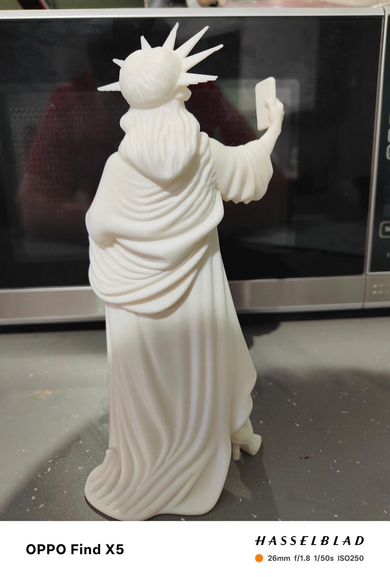K&ouml;nnte beinhalten: Eine wei&szlig;e Statue der Freiheitsstatue, die ein Selfie mit einem Smartphone macht. Die Statue tr&auml;gt eine stachelige Krone und drapierte Gew&auml;nder. Das Bild ist gut beleuchtet und hebt die Details der Skulptur hervor.