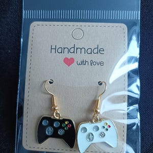 May include: A pair of handmade earrings featuring a black and a white video game controller design. The controllers have colorful buttons and are attached to gold-colored fish hook ear wires. The earrings are displayed on a tan card with the text "Handmade with love."