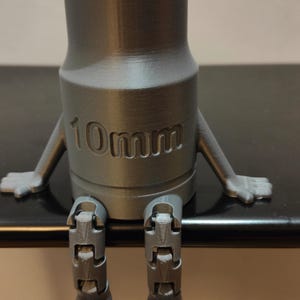 May include: A gray 3D-printed socket wrench with arms and legs, sitting down. The wrench is labeled "10mm" and has a cartoonish design. The object is on a dark surface.