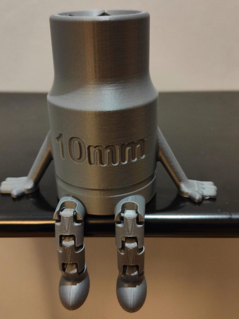 May include: A gray 3D-printed socket wrench with arms and legs, sitting down. The wrench is labeled "10mm" and has a cartoonish design. The object is on a dark surface.