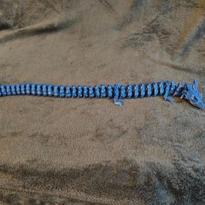 May include: A blue articulated dragon toy, lying on a gray surface. The dragon is made of many small, connected pieces, allowing it to move and pose. The dragon's head and tail are detailed with sharp features.