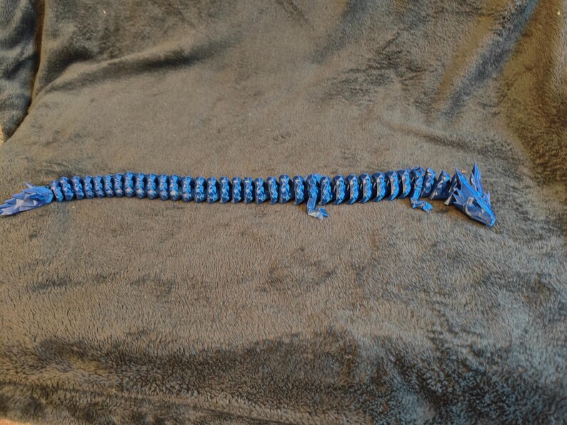 May include: A blue articulated dragon toy, lying on a gray surface. The dragon is made of many small, connected pieces, allowing it to move and pose. The dragon's head and tail are detailed with sharp features.