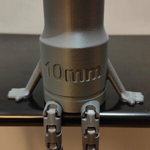 May include: A gray, 3D-printed socket wrench with arms and legs, giving it a cartoon-like appearance. The text "10mm" is embossed on the wrench. The wrench is sitting on a dark surface.