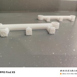 May include: Two white, 3D-printed, bar-shaped objects with cylindrical ends and square, textured protrusions. The objects are positioned on a speckled gray surface, with one in the foreground and another in the background.