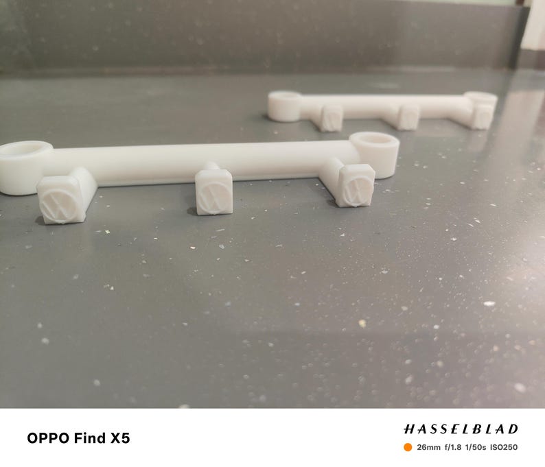 May include: Two white, 3D-printed, bar-shaped objects with cylindrical ends and square, textured protrusions. The objects are positioned on a speckled gray surface, with one in the foreground and another in the background.