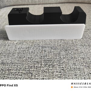 以下が含まれることがあります： 白と黒の3Dプリントされた電話スタンド。白い長方形のベースと、湾曲した切り欠きのある黒い上部セクションがあります。「OPPO Find X5」の文字が下部に表示されています。