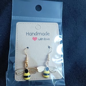 May include: Bee-shaped earrings with gold hooks and wings. The bee bodies are yellow with black stripes. The earrings are presented on a small, brown card with the text "Handmade with love."