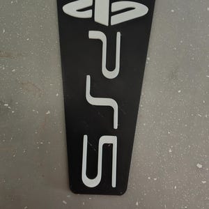 May include: Black and white PS5 logo and controller button symbols on a black background. The PS5 logo is in white and is the primary focus of the image. The background is a dark gray color.