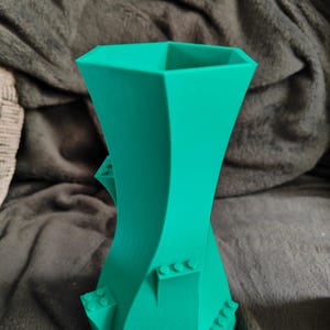 May include: A teal-colored, 3D-printed vase with a unique, twisted design. The vase features a geometric shape with integrated block-like details, adding a playful touch to its modern aesthetic. The vase is suitable for home decor.