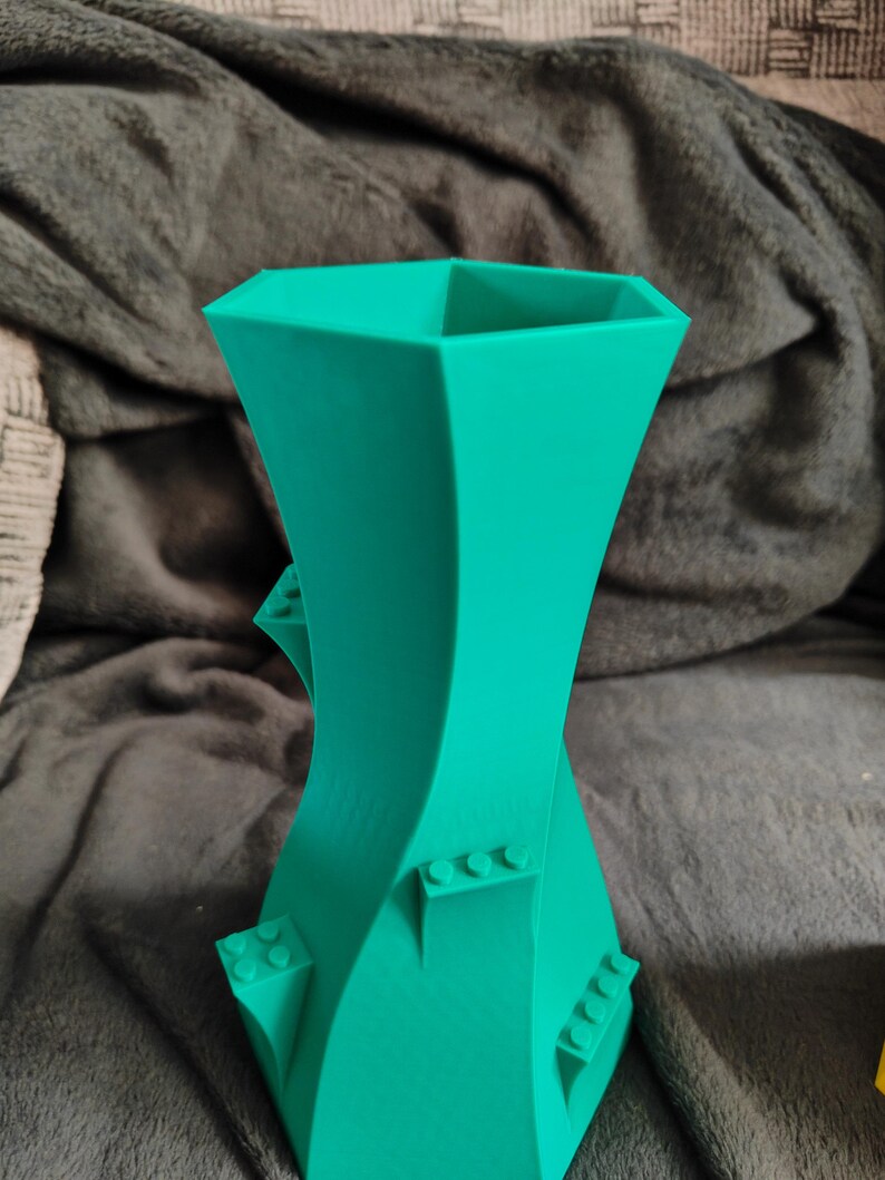 May include: A teal-colored, 3D-printed vase with a unique, twisted design. The vase features a geometric shape with integrated block-like details, adding a playful touch to its modern aesthetic. The vase is suitable for home decor.