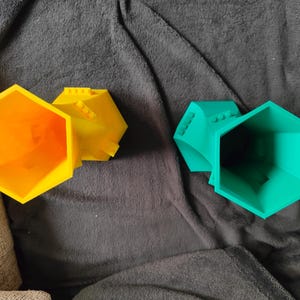 May include: Three colorful, geometric, hexagonal containers. One is bright orange, another is yellow, and the third is teal. The containers have a Lego-like design on the top. The objects are on a dark gray, textured surface.