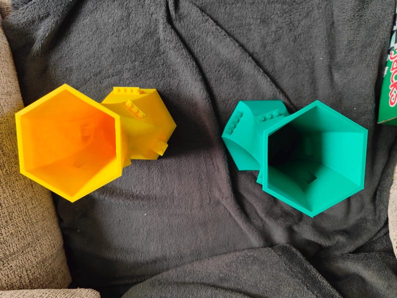 May include: Three colorful, geometric, hexagonal containers. One is bright orange, another is yellow, and the third is teal. The containers have a Lego-like design on the top. The objects are on a dark gray, textured surface.