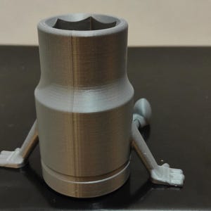 May include: A gray, 3D-printed socket wrench with arms and hands. The wrench has a hexagonal opening and a cylindrical body. The arms are extended, and the hands are open.