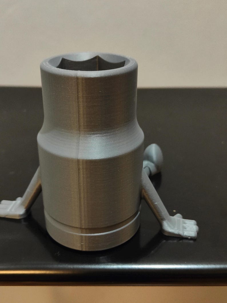 May include: A gray, 3D-printed socket wrench with arms and hands. The wrench has a hexagonal opening and a cylindrical body. The arms are extended, and the hands are open.