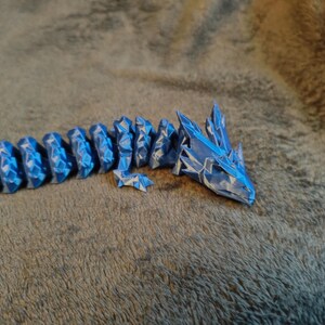 May include: A blue and white articulated dragon toy. The dragon is made of segments that allow it to be posed. The head and body are detailed with a geometric, crystalline design. The toy is resting on a textured, gray surface.