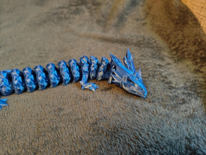 May include: A blue and white articulated dragon toy. The dragon is made of segments that allow it to be posed. The head and body are detailed with a geometric, crystalline design. The toy is resting on a textured, gray surface.