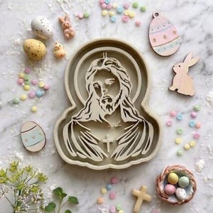 May include: An Easter-themed arrangement featuring a Jesus-shaped cookie cutter, wooden Easter egg and bunny cutouts, and pastel-colored candies. Decorative eggs and a small nest with eggs are also present, all arranged on a white surface.