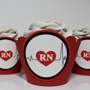 May include: Three cupcakes with gray frosting sit in red cupcake wrappers. The wrappers feature a white circle with a red heart, the letters "RN", and a heartbeat line. The cupcakes are on a white surface.